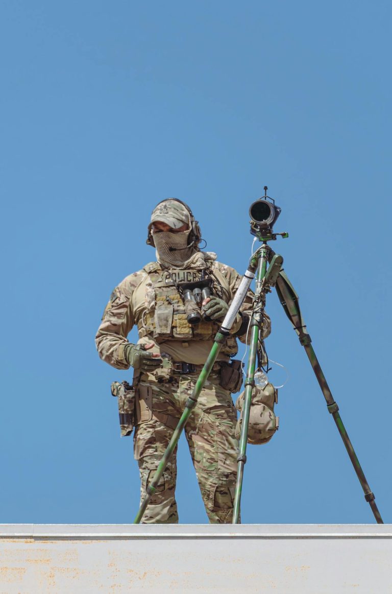 Armed military personnel in camo gear on roof, overlooking airshow event for security.