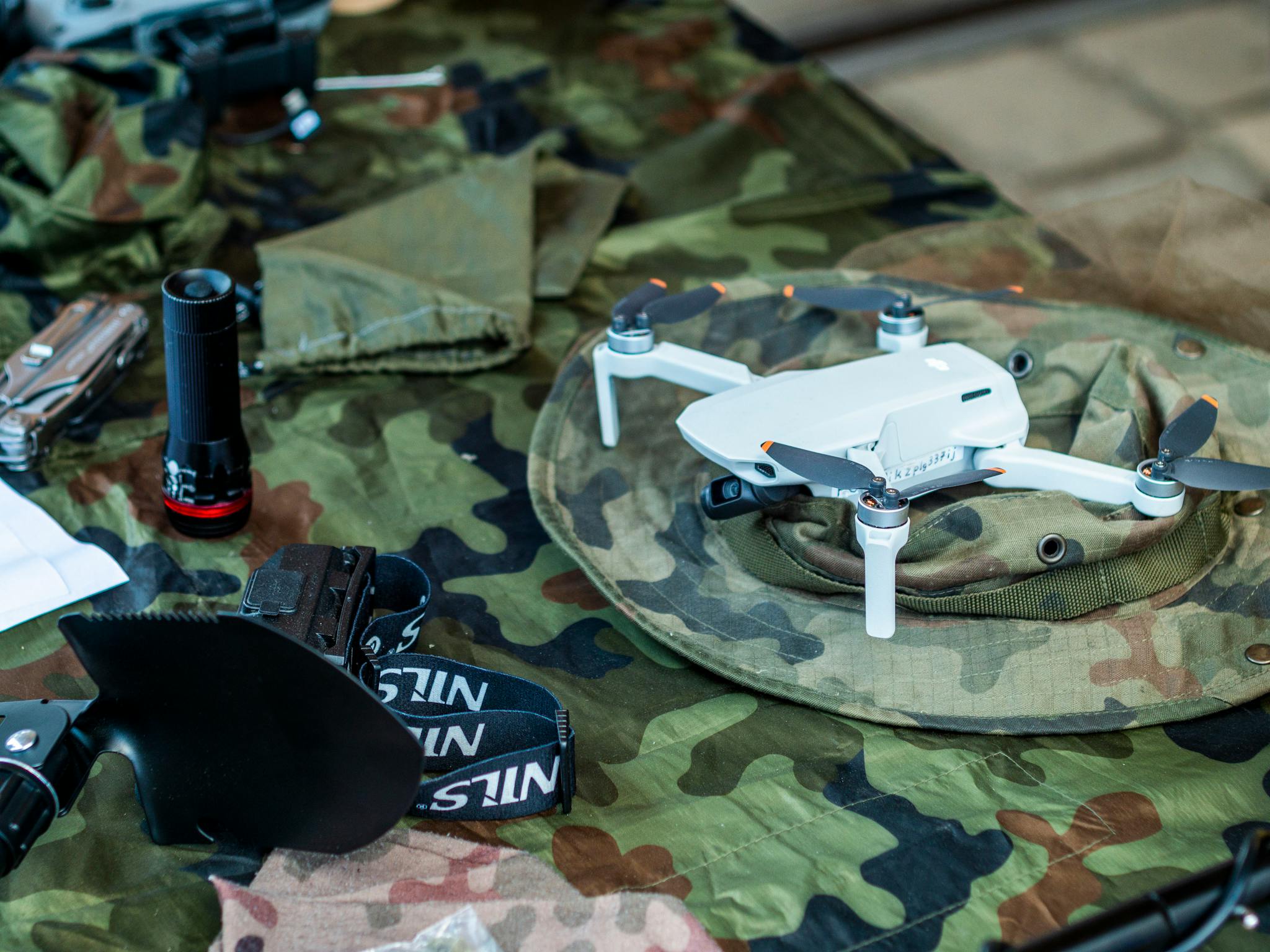 Compact white drone on camouflage gear, surrounded by flashlight and tools.
