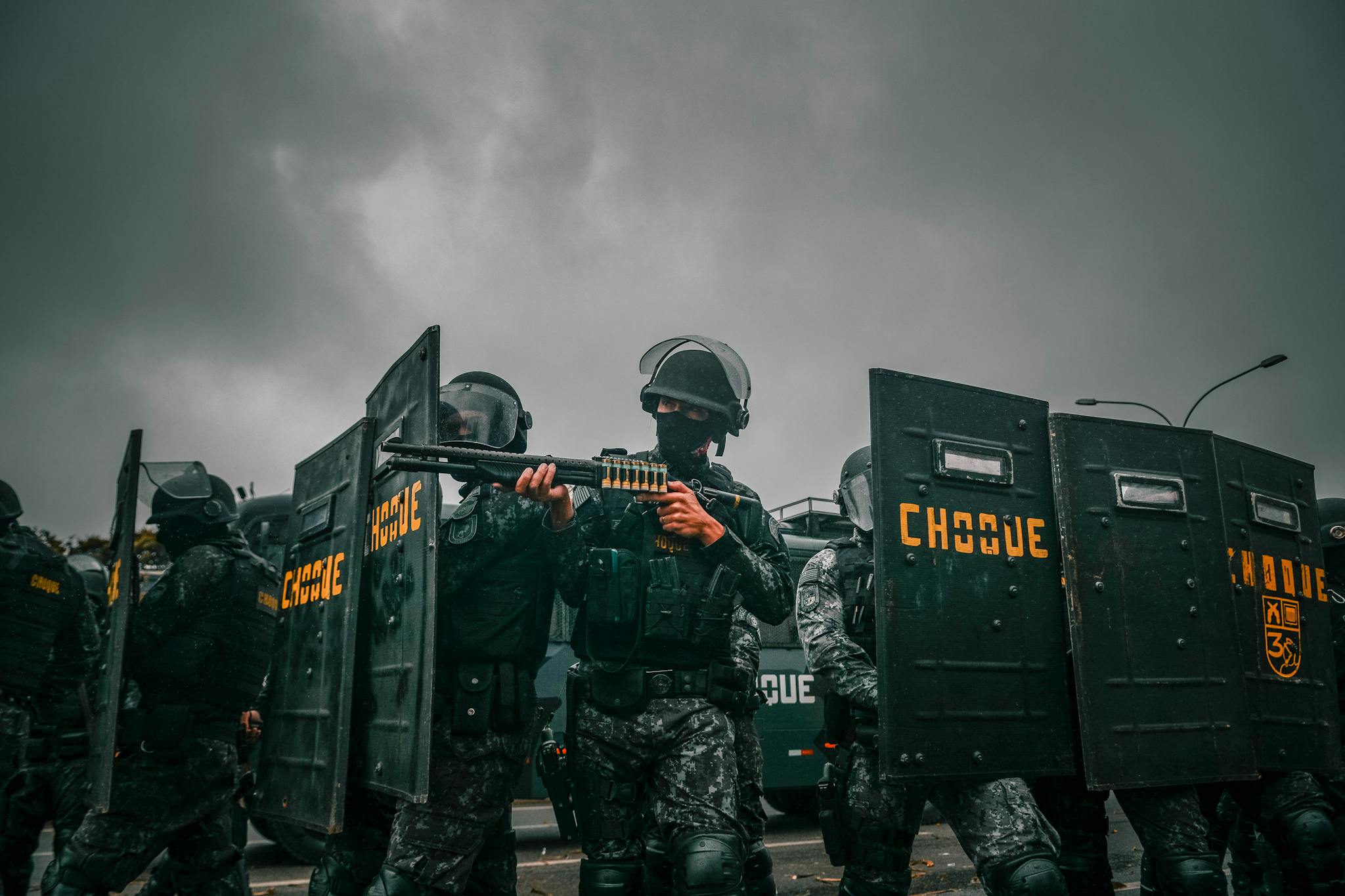 Riot police in tactical gear holding shields during a stormy protest.