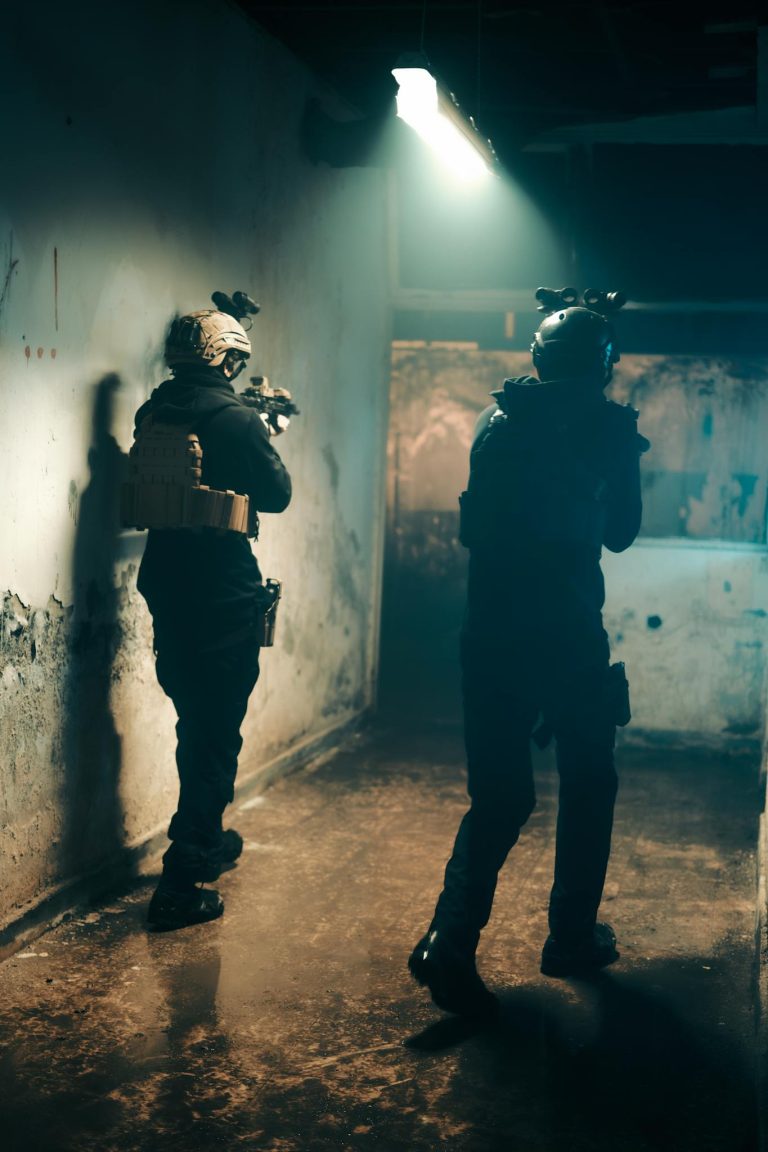 Two military operatives in tactical gear navigate a dimly lit indoor environment.